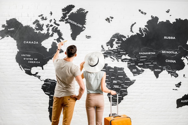 Молодая пара путешественников, стоящих рядом с большой картой мира на заднем плане, выбирая место для летних каникул
