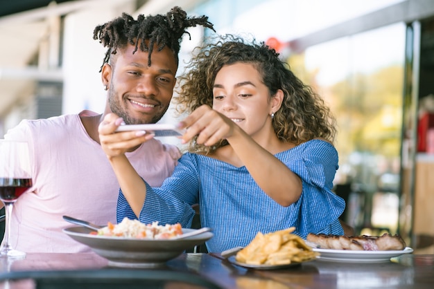 写真 携帯電話を使って、レストランで一緒に昼食をとりながら食事の写真を撮る若いカップル。