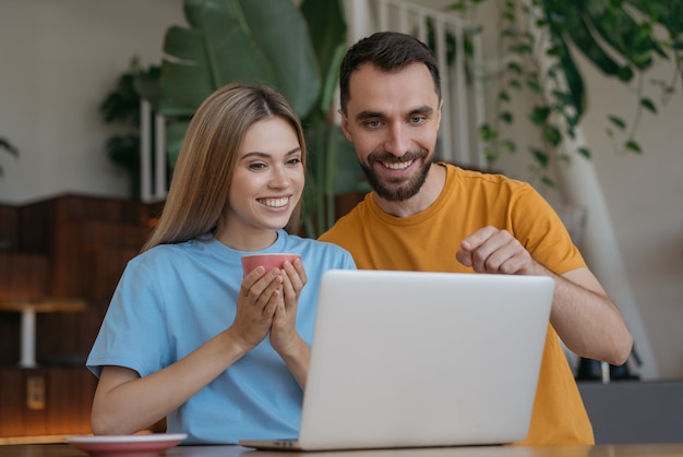 Молодая пара, используя ноутбук, делая покупки в Интернете дома. Улыбающийся мужчина и женщина делают видеоконференцсвязь