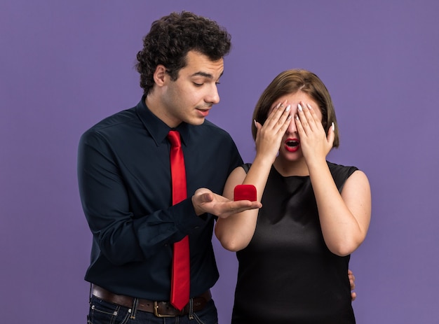 Young couple on valentine's day excited man giving engagement ring to woman looking at ring curious woman covering eyes with hands isolated on purple wall