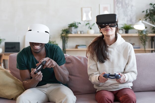 Young couple in virtual reality glasses sitting on sofa and playing video game in team in the room