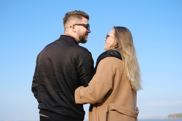 A young couple in warm outerwear stands against the blue sky Man and woman look at each other and smile