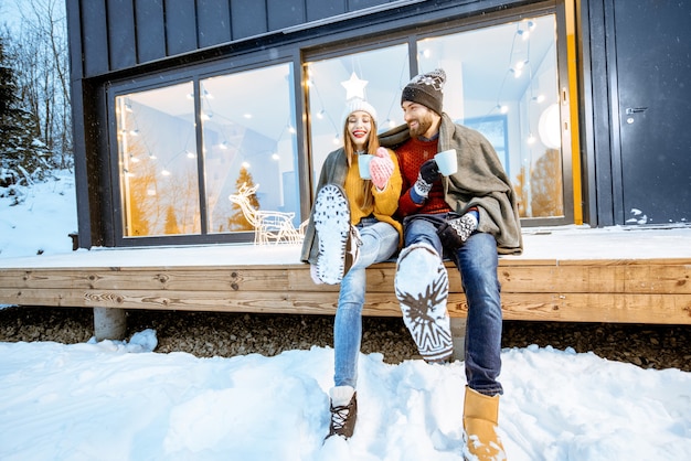 Young couple warming up with plaid and hot drinks sitting on the terrace of the modern house in the mountains durnig the winter holidays