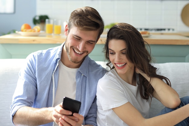 Young couple watching online content in a smart phone sitting on a sofa at home in the living room.