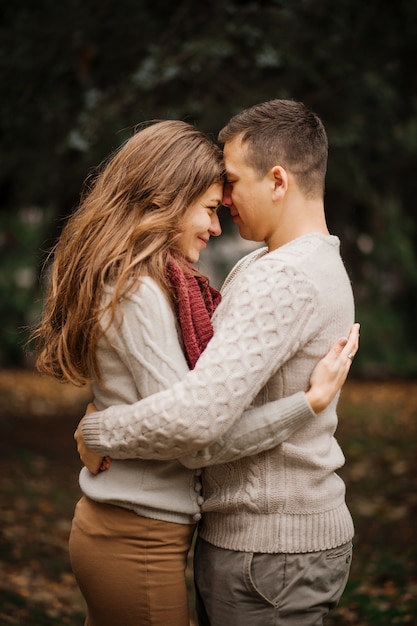 Young couple wearing on tied warm sweaters hugging in love at city in autumn background yellow bushes and trees.