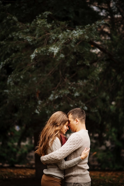 Young couple wearing on tied warm sweaters hugging in love at city in autumn background yellow bushes and trees.