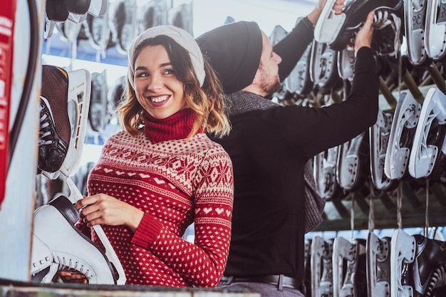 Young couple wearing warm clothes standing near rack with many pairs of skates, choosing his size