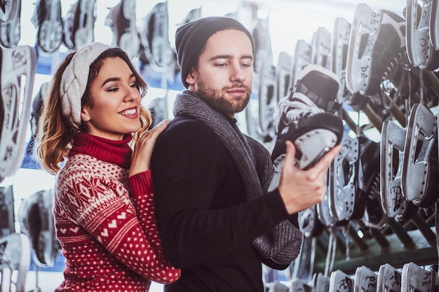 Young couple wearing warm clothes standing near rack with many pairs of skates, choosing his size