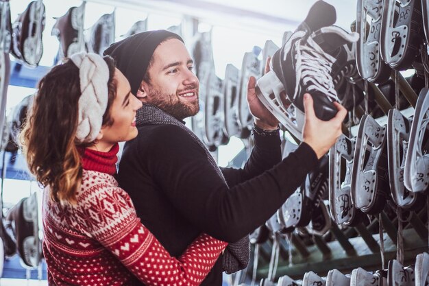 Young couple wearing warm clothes standing near rack with many pairs of skates, choosing his size