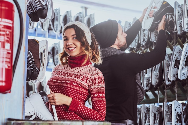 Young couple wearing warm clothes standing near rack with many pairs of skates, choosing his size