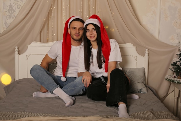 흰색 티셔츠와 산타클로스 모자를 쓴 젊은 부부가 침대에 앉아 있다