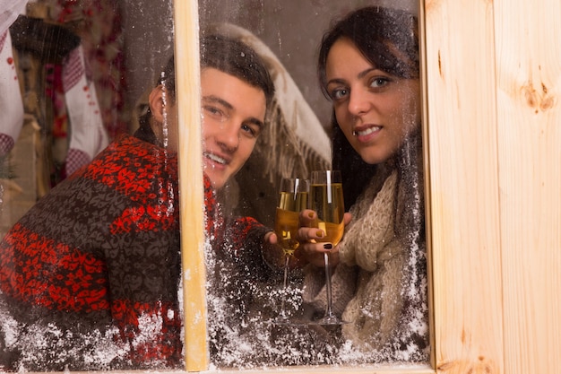 Молодая пара в зимних нарядах держит бокалы шампанского за прозрачным стеклянным окном, улыбаясь в камеру