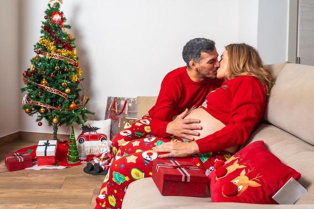 装飾と赤いクリスマス服を着た若いカップルがお腹の赤ちゃんの隆起に触れ、キスをする妊娠中の女性の家族