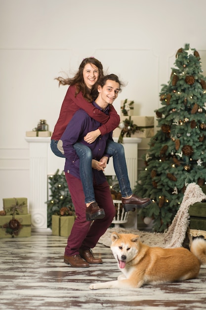 A young couple with a dog fooling around near a Christmas tree