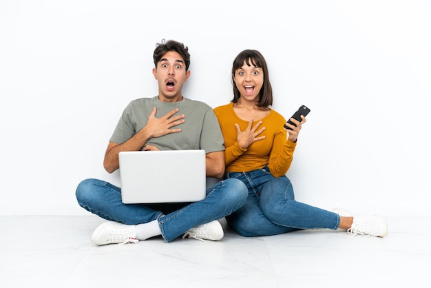 Young couple with a laptop and mobile sitting on the floor surprised and shocked while looking right