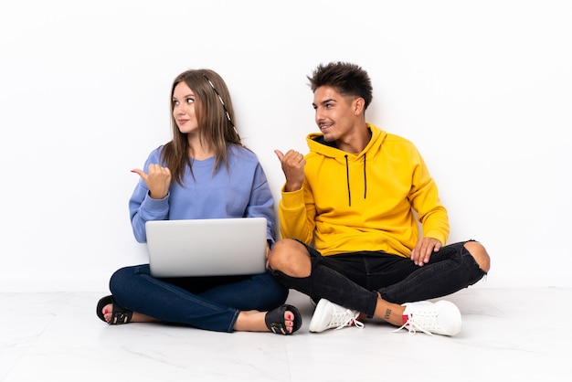 Молодая пара с ноутбуком, сидя на полу на белом, указывая в сторону, чтобы представить продукт