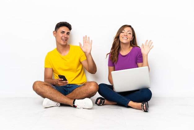 Giovani coppie con un computer portatile che si siede sul pavimento su bianco che saluta con la mano con l'espressione felice