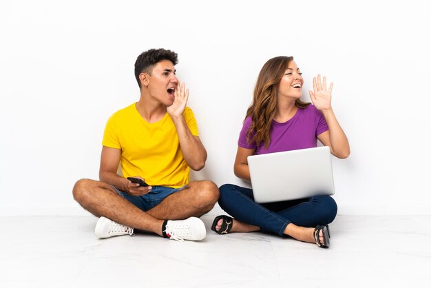 Young couple with a laptop sitting on the floor on white shouting with mouth wide open to the side