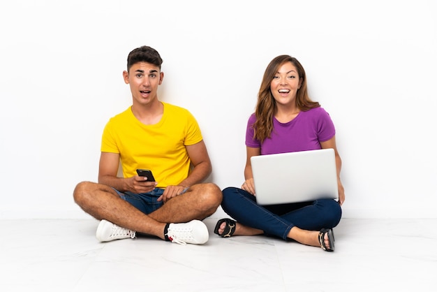 Young couple with a laptop sitting on the floor on white with surprise facial expression