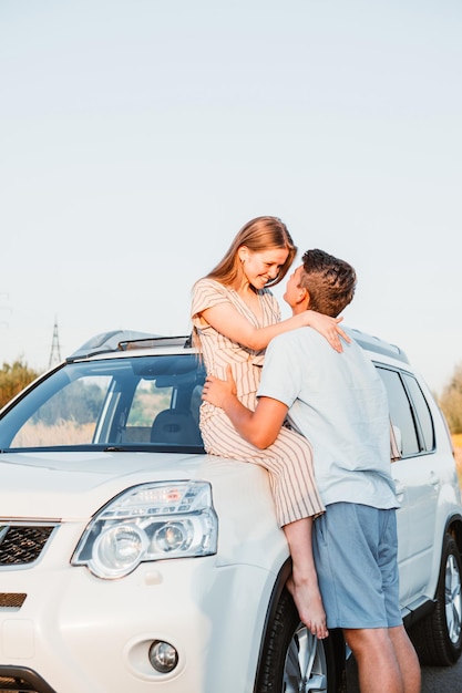 Young couple woman sitting on car hood man kissing her. copy space