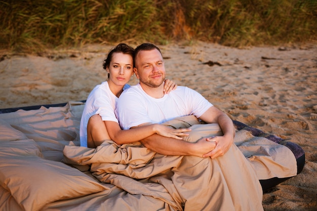 Young couple wrapped in a blanket outdoors happy couple in love lying and rotating in bed by the sea at sunset happy caucasian couple of husband and wife wrapped in a blanket outdoors