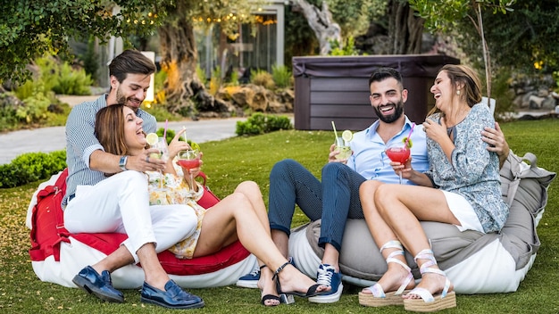 Young couples having fun in the meadow of a resort luxury holidays for millennials happy people laughing and drinking sitting on garden sofas