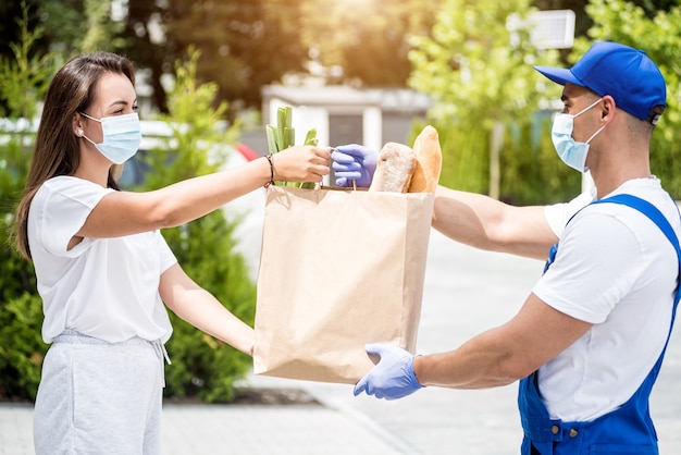 Il giovane corriere che indossa una maschera protettiva e guanti consegna merci a una giovane donna durante la quarantena