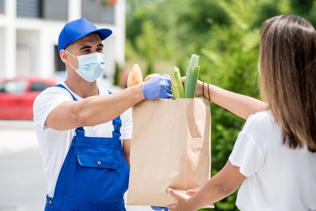 Il giovane corriere che indossa una maschera protettiva e guanti consegna merci a una giovane donna durante la quarantena.