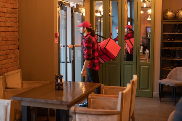 Молодой продавец с большой красной сумкой на плече собирается открыть дверь кафе, уходя доставлять заказы клиентам