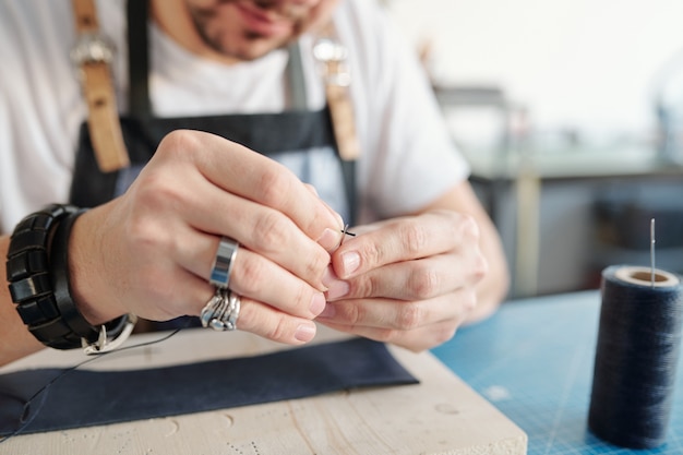 Young craftsman putting black thread into needle hole over wooden board with piece of leather before sewing