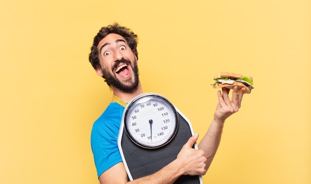 Espressione felice del giovane atleta barbuto pazzo, tenendo panino e bilancia