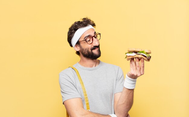 Giovane atleta barbuto pazzo che pensa espressione e tiene un panino