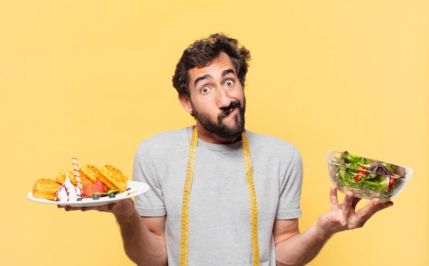 Young crazy bearded man dieting doubting or uncertain expression and holding a salad and waffles