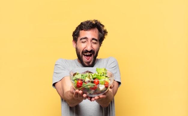 Giovane uomo barbuto pazzo che è a dieta espressione felice e tiene un'insalata