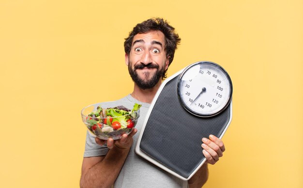 Giovane uomo barbuto pazzo che è a dieta espressione felice e tiene in mano una bilancia e un'insalata