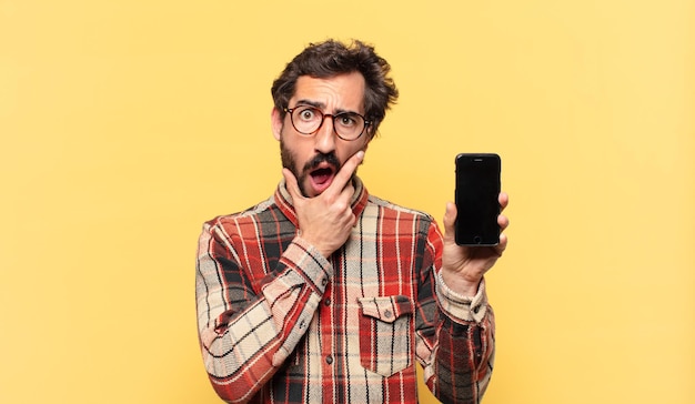 Молодой сумасшедший бородатый мужчина сомневается или неуверенно смотрит в камеру и держит телефон