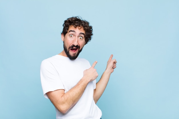 Young crazy bearded man feeling joyful and surprised, smiling with a shocked expression and pointing to the side