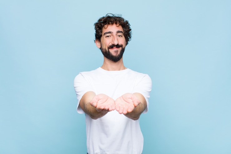 Photo young crazy bearded man smiling happily with friendly, confident, positive look, offering and showing an object or concept on flat wall