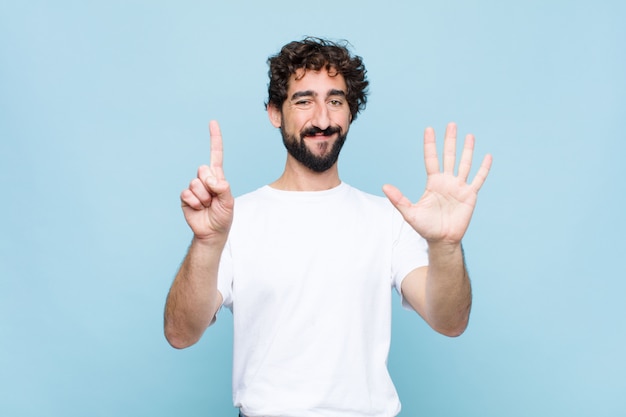 Young crazy bearded man smiling and looking friendly, showing number six or sixth with hand forward, counting down on flat wall