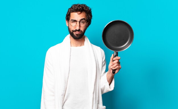 Young crazy bearded man wearing a bathrobe cooking with a pan