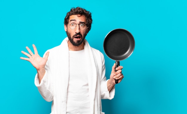 Young crazy bearded man wearing a bathrobe cooking with a pan