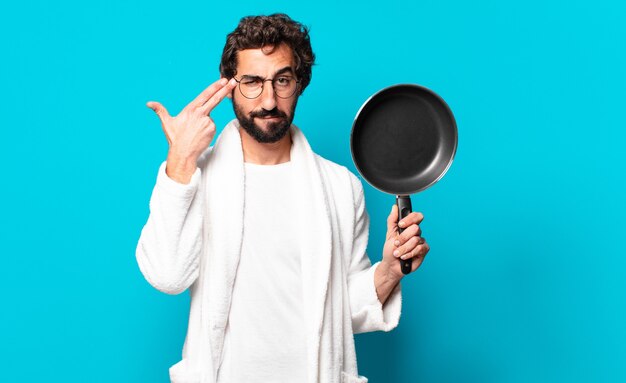 Young crazy bearded man wearing a bathrobe cooking with a pan