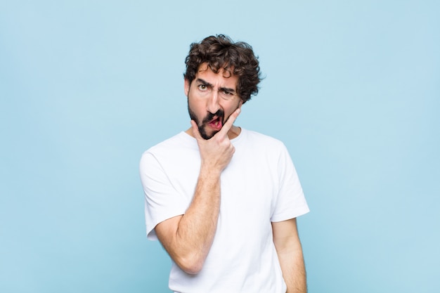 Young crazy bearded man with mouth and eyes wide open and hand on chin, feeling unpleasantly shocked, saying what or wow over wall