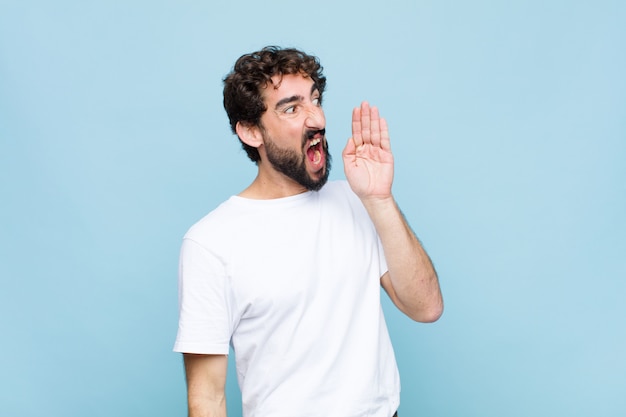 Young crazy bearded man yelling loudly and angrily to copy space on the side, with hand next to mouth over wall