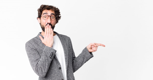 Young crazy businessman feeling happy, shocked and surprised, covering mouth with hand and pointing to lateral copy space against flat wall