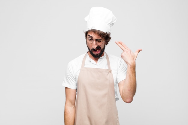 Young crazy chef looking unhappy and stressed, suicide gesture making gun sign with hand, pointing to head against white wall