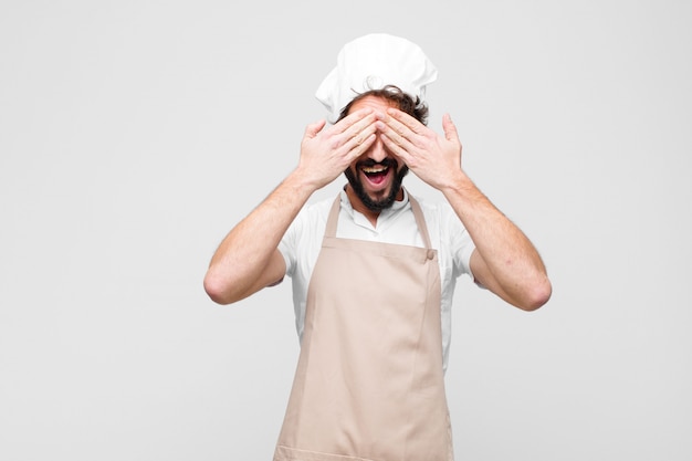 Young crazy chef smiling and feeling happy, covering eyes with both hands and waiting for unbelievable surprise against white wall