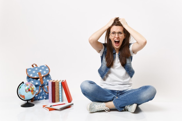 Young crazy dizzy woman student in denim clothes screaming clinging to head sitting near globe, backpack school books isolated