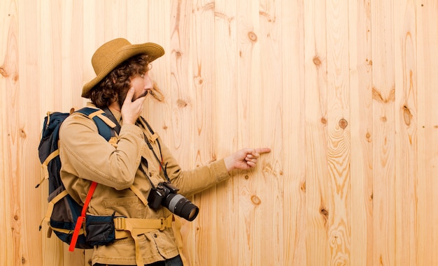 Young crazy explorer with straw hat and backpack on wooden wall 