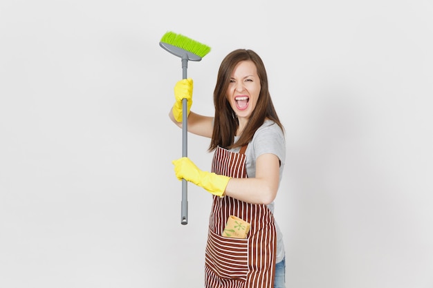 縞模様のエプロン、白い背景で隔離の黄色い手袋の若い狂気の主婦。ほうきを持って掃除するメイドを掃除する楽しい家政婦の女性。広告用のスペースをコピーします。広告エリア。